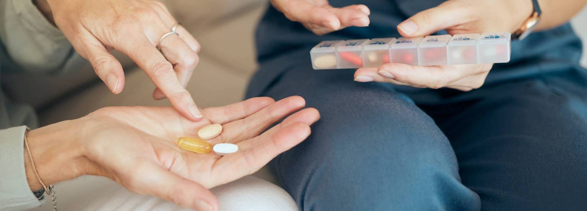 Two people reviewing pills from a weekly pill organizer, with three tablets held in an open palm.